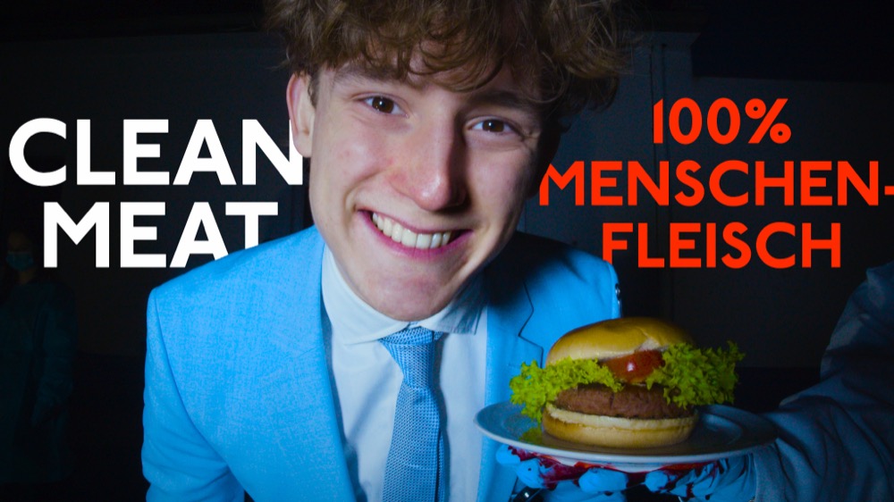 Szene aus dem Kurzfilm „Clean Meat – 100% Menschlich“: Ein lächelnder Mann im hellblauen Anzug serviert einen Burger, daneben der Schriftzug „100% Menschenfleisch“